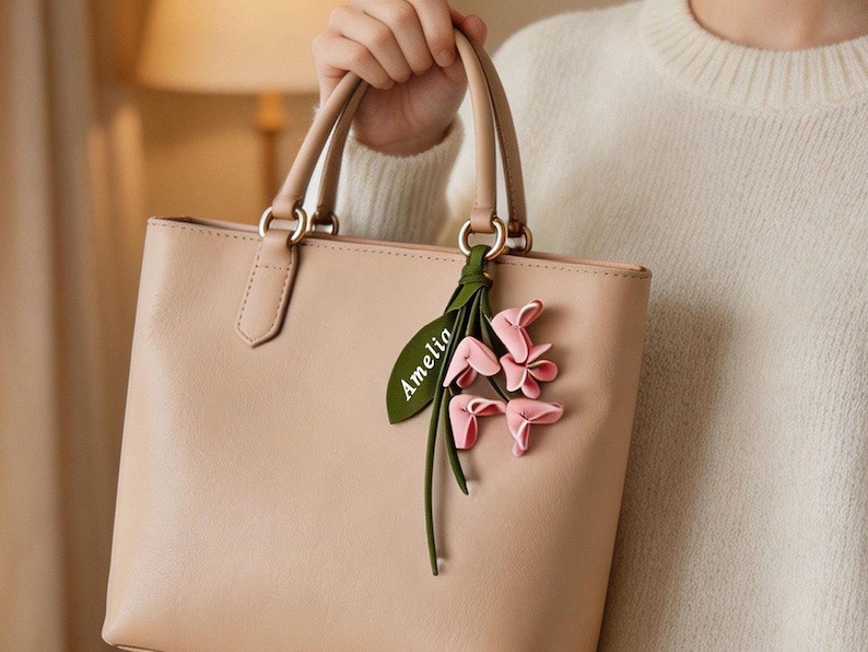 May include: A beige leather handbag with two top handles, featuring a decorative green leaf-shaped charm with the name Amelia printed in white, accompanied by a cluster of small, soft pink fabric flowers attached to the handle ring.