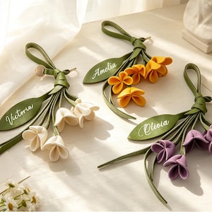 May include: Three decorative bag charms featuring green leather-like straps and leaves with personalized names Victoria, Amelia, and Olivia. Each charm includes a cluster of three fabric flowers in white, yellow, and purple, attached to a loop.