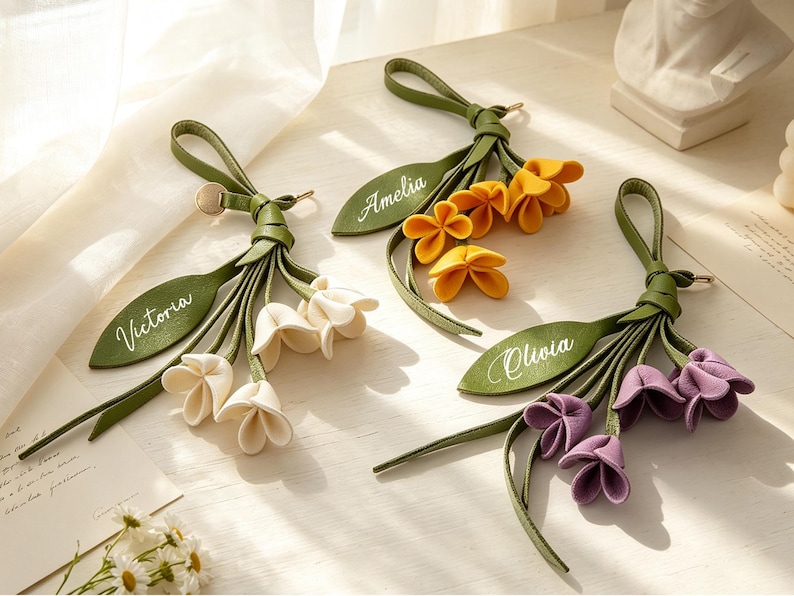 May include: Three decorative bag charms featuring green leather-like straps and leaves with personalized names Victoria, Amelia, and Olivia. Each charm includes a cluster of three fabric flowers in white, yellow, and purple, attached to a loop.