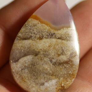 Pode incluir: Um cabochão de ágata em forma de lágrima com um padrão em camadas de marrom, branco e roxo. A pedra tem uma superfície lisa e polida.