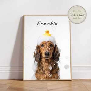 May include: A framed print featuring a long-haired dachshund named Frankie, adorned with a foam bath and a yellow rubber ducky on its head. The print has a light wood frame and includes the text "Frankie" and "Designed by Debra Gail Barn & Brush."