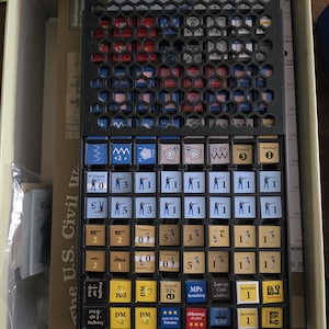 May include: A board game with a black plastic organizer filled with small square game pieces. The pieces are various colors, including red, blue, yellow, and brown, and feature numbers, symbols, and flags. The game appears to be a historical strategy game.