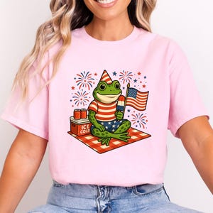 May include: Pink t-shirt featuring a cartoon frog celebrating Independence Day. The frog wears a patriotic hat, striped shirt, and star-spangled shorts, holding a popsicle and an American flag. A cooler with "ICE COLD" text and fireworks complete the design.