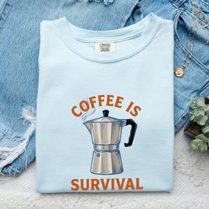Puede incluir: Una camiseta de cuello redondo en color azul claro con una etiqueta de Comfort Colors en el interior del cuello. El frente presenta un gráfico de una cafetera italiana plateada con asa negra, junto al texto naranja "COFFEE IS" arriba y "SURVIVAL" abajo.