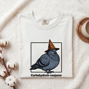 May include: A white t-shirt featuring a graphic of a grey pigeon wearing an orange wizard hat with yellow stars inside a black square frame. Below the frame, the text reads Carbohydrate conjurer in a bold, black, sans-serif font.