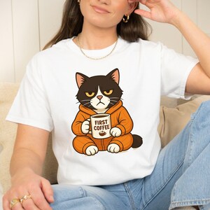 Könnte beinhalten: Weißes T-Shirt mit einer Comic-Katze in einem orangefarbenen Overall, die eine Kaffeetasse hält. Auf der Tasse steht "FIRST COFFEE". Die Katze hat einen grimmigen Gesichtsausdruck, schwarz-braunes Fell und gelbe Augen.