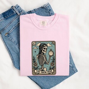 May include: A light pink t-shirt featuring a rectangular graphic of a skeleton wearing goggles and holding a stopwatch, framed by flowers and clouds. The graphic includes the Roman numeral VI at the top and the text THE SWIM MOM at the bottom.