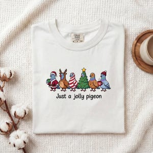 May include: A white t-shirt featuring a graphic of six pigeons dressed in holiday attire, including a Santa suit, reindeer antlers, a candy cane pattern, a Christmas tree, and winter hats. The text below the pigeons reads Just a jolly pigeon.