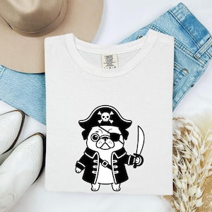 May include: A white short-sleeved t-shirt featuring a black graphic of a pug dressed as a pirate, wearing an eye patch and a hat with a skull and crossbones, holding a sword. The inner neck label displays the text Comfort Colors L-G-G.