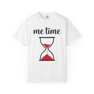 May include: A white short-sleeve t-shirt featuring the black text "me time" printed above a graphic of an hourglass. The hourglass has a black outline and contains red sand, with some grains depicted falling from the top bulb to the bottom.
