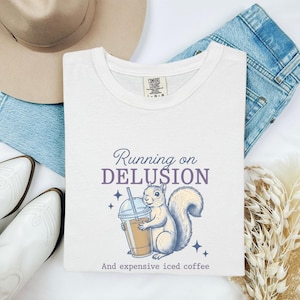 May include: A white short-sleeve t-shirt featuring a graphic of a squirrel holding an iced coffee cup. The text reads Running on DELUSION And expensive iced coffee. The design includes small star accents in blue and purple ink on the fabric.