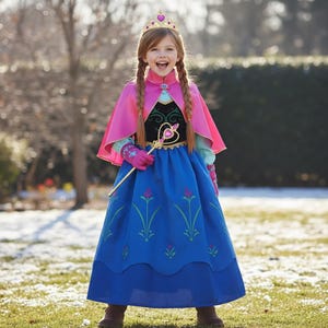 Peut inclure: Une jeune fille porte un costume de princesse, avec une cape rose, une robe bleue et une couronne dorée. La robe présente des motifs floraux. Elle tient une baguette dorée avec un cœur. L'arrière-plan est une scène extérieure enneigée.