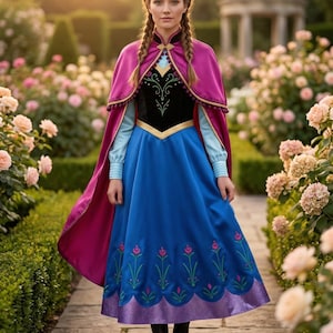 Peut inclure: Une femme porte une robe bleue et noire avec une cape rose sur un chemin de pierre dans un jardin. La robe a des broderies florales et une bordure dorée. La cape est rose foncé. Le fond présente des rosiers et un belvédère.