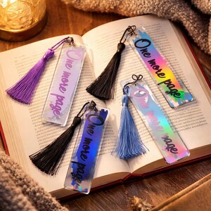 Peut inclure: Plusieurs marque-pages en acrylique transparent avec des pompons colorés. Chaque marque-page porte l'inscription "One more page". Les pompons sont de couleurs variées, dont le violet, le noir et le bleu. Les marque-pages sont posés sur un livre ouvert.
