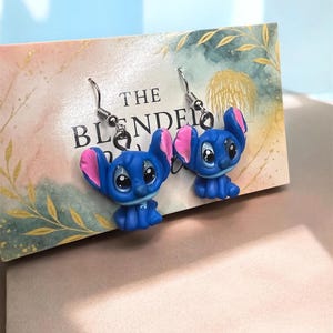 May include: A pair of blue Stitch earrings with pink inner ears and silver hooks. The earrings are displayed on a decorative card with the text "THE BLENDED". The background is a soft watercolor design.