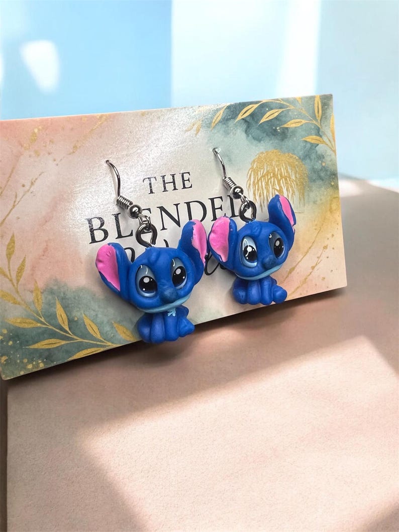 May include: A pair of blue Stitch earrings with pink inner ears and silver hooks. The earrings are displayed on a decorative card with the text "THE BLENDED". The background is a soft watercolor design.