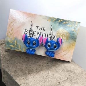 May include: A pair of blue and pink character earrings with silver hooks. The earrings are displayed on a card with the text "THE BLENDED". The background is a light watercolor design.