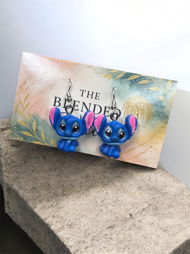 May include: A pair of blue and pink character earrings with silver hooks. The earrings are displayed on a card with the text "THE BLENDED". The background is a light watercolor design.