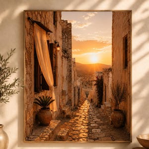 May include: A rectangular framed wall art print depicting a narrow stone alleyway in a Mediterranean village at sunset. The scene features golden light, stone buildings, a sheer curtain in a window, potted plants, and a cobblestone path.