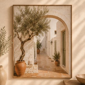 May include: A framed wall art print depicting a Mediterranean stone alleyway with a large olive tree in a terracotta pot in the foreground. The scene features white stone walls, arched doorways, green wooden doors, and a tiled path under warm sunlight.