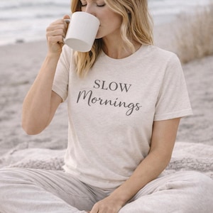 May include: A person wearing a light beige t-shirt and light grey trousers, sitting on a blanket, drinking from a white mug. The t-shirt has the words "SLOW Mornings" printed on it in a dark grey script.