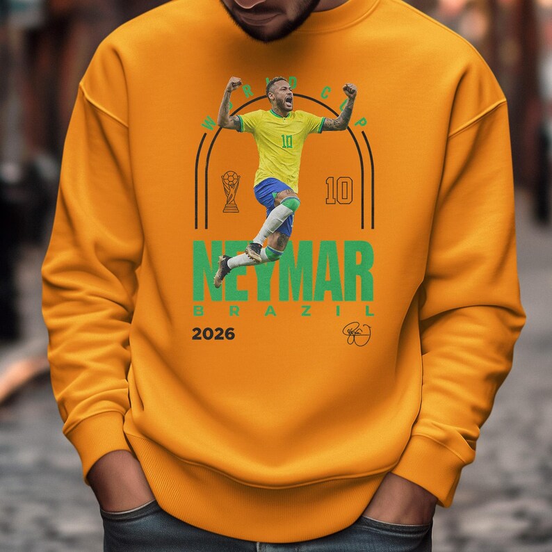 A man wearing a yellow sweatshirt with a graphic of a soccer player in mid-jump, celebrating.