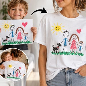 Könnte beinhalten: Ein weißes T-Shirt mit einer bunten Kinderzeichnung aus Wachsmalstiften, die eine Familie mit Sonne, Herz, Hund und drei Figuren auf grünem Gras zeigt. Das Motiv ist vorne aufgedruckt, ein kleinerer kreisförmiger Ausschnitt zeigt die Rückseite.
