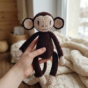 May include: A crocheted monkey toy made of dark brown and cream-colored yarn. The monkey features a round head with large circular ears, a small embroidered face, long slender arms and legs, and cream-colored hands and feet.