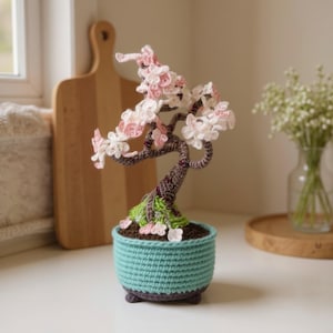 May include: A crocheted bonsai tree featuring a textured brown trunk and branches adorned with delicate pink and white flowers. The tree is rooted in a patch of green yarn moss and sits inside a round, light blue crocheted pot with small feet.