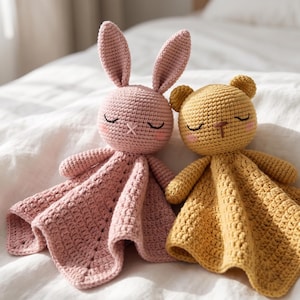 May include: Two handmade crochet security blankets featuring a pink rabbit and a yellow bear. Both plush toys have closed eyes, stitched noses, and soft pink cheeks. The bodies consist of a textured, square-shaped blanket with a crocheted finish.