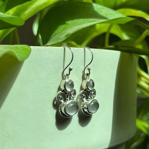 Peut inclure: Une paire de boucles d'oreilles pendantes en argent avec deux pierres rondes, translucides et bleu pâle serties dans du métal poli. Chaque boucle possède un crochet incurvé et des volutes décoratives reliant les deux pierres en serti clos.