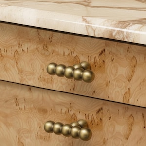 May include: Close-up of a wooden dresser with two drawers. The drawers have a light wood grain finish and are adorned with brass-coloured, spherical drawer pulls. The top of the dresser is a light-coloured marble.