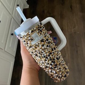 May include: A bedazzled Stanley tumbler with a white handle, lid, and straw. The tumbler is covered in a pattern of black, white, and gold beads. The Stanley logo is visible on the front.