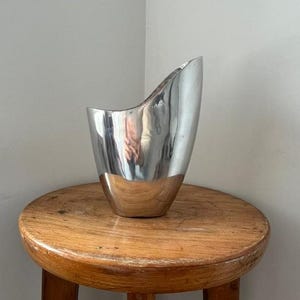 Pu&ograve; includere: Un vaso d'argento lucido con un design unico e asimmetrico. Il vaso ha una base ampia e arrotondata che si assottiglia verso l'alto fino a una parte superiore curva e aperta. &Egrave; posto su uno sgabello rotondo di legno.