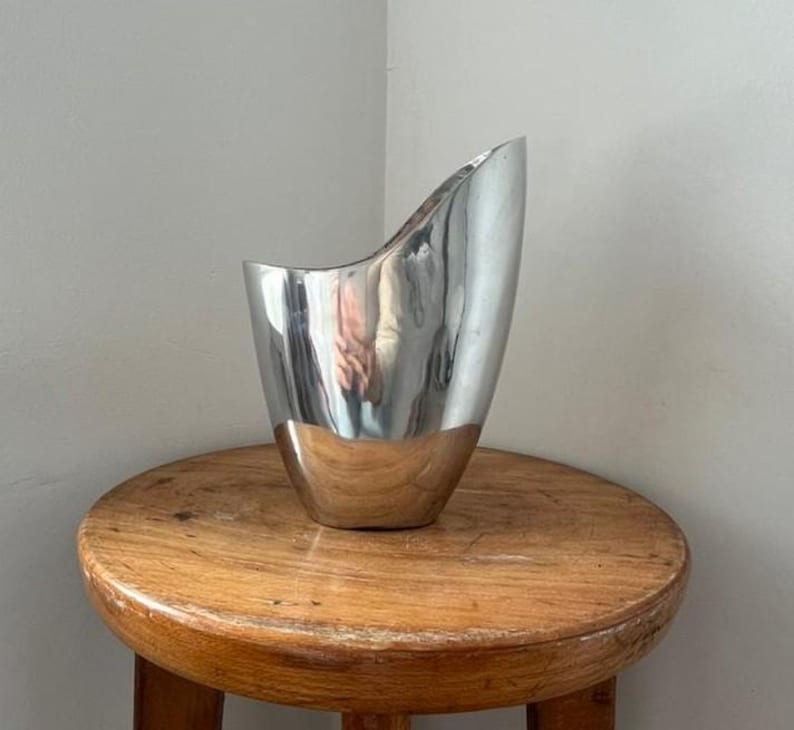Pu&ograve; includere: Un vaso d'argento lucido con un design unico e asimmetrico. Il vaso ha una base ampia e arrotondata che si assottiglia verso l'alto fino a una parte superiore curva e aperta. &Egrave; posto su uno sgabello rotondo di legno.