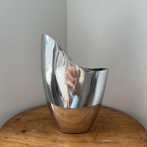 Pu&ograve; includere: Un moderno vaso in metallo argentato con un design asimmetrico unico. Il vaso ha una superficie lucida e riflettente e una base in metallo spazzolato a contrasto. Il vaso &egrave; esposto su un tavolo rotondo di legno.