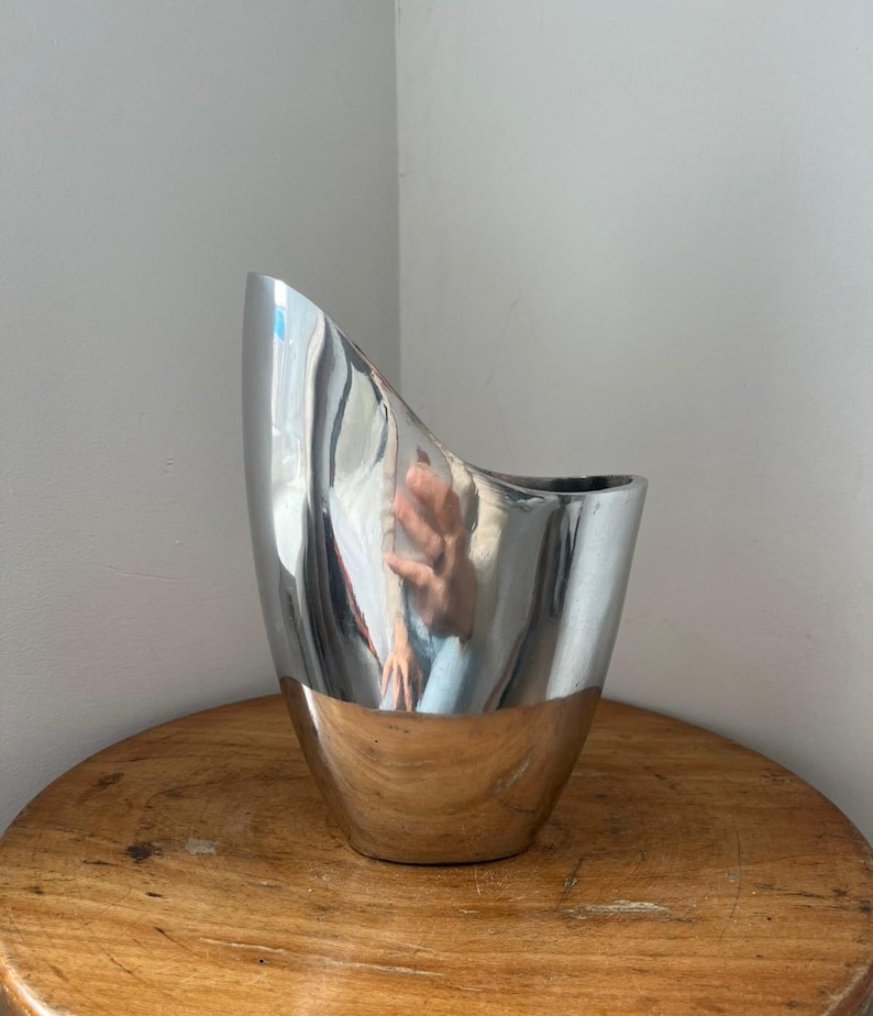 Pu&ograve; includere: Un moderno vaso in metallo argentato con un design asimmetrico unico. Il vaso ha una superficie lucida e riflettente e una base in metallo spazzolato a contrasto. Il vaso &egrave; esposto su un tavolo rotondo di legno.