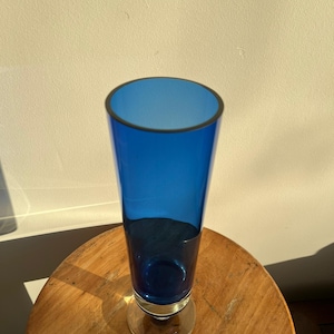 Op de afbeelding: Een hoge, kobaltblauwe glazen vaas met een heldere, cirkelvormige basis en een donkere rand. De vaas is cilindrisch, met een licht uitlopende opening. Hij staat op een ronde, houten tafel.