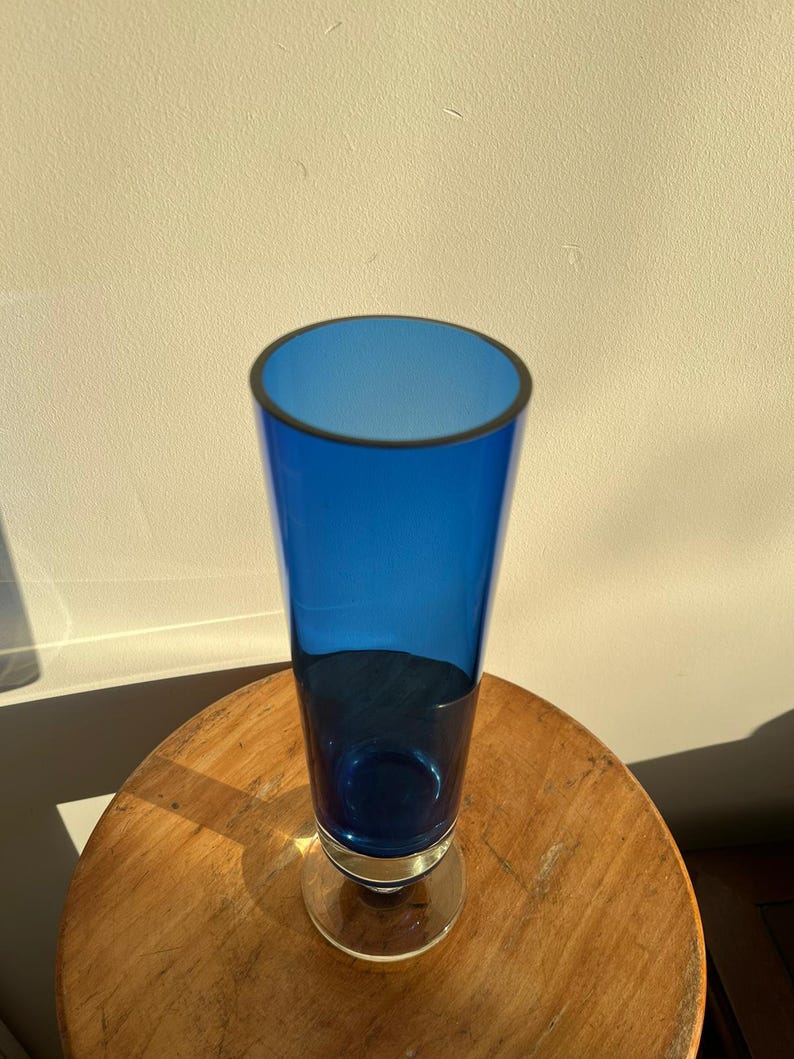 Op de afbeelding: Een hoge, kobaltblauwe glazen vaas met een heldere, cirkelvormige basis en een donkere rand. De vaas is cilindrisch, met een licht uitlopende opening. Hij staat op een ronde, houten tafel.