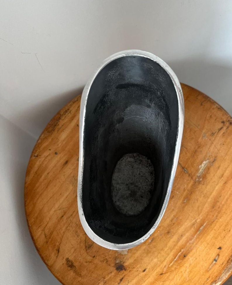 Pu&ograve; includere: Un contenitore argentato di forma ovale con un interno scuro. Il contenitore ha una finitura metallica liscia all'esterno e poggia su una superficie di legno. L'interno sembra essere di una tonalit&agrave; pi&ugrave; scura, forse nera.