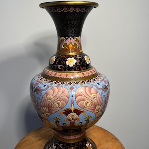 May include: A decorative vase featuring a complex cloisonné-style pattern with a black neck and base, accented by white floral motifs. The bulbous body displays a light blue background with symmetrical pink and orange fan-shaped designs.