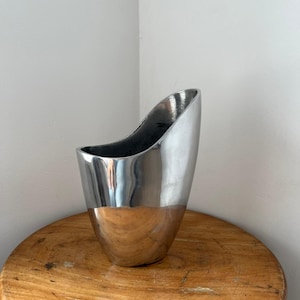Pu&ograve; includere: Un vaso in metallo argentato lucido con un design unico e asimmetrico. Il vaso ha un'ampia apertura e una base arrotondata. La superficie riflettente del vaso cattura la luce, creando riflessi e ombre. Misura circa 25 cm di altezza.