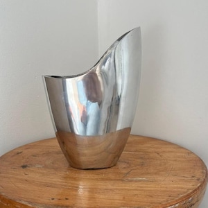 Pu&ograve; includere: Un vaso argentato lucido con un design asimmetrico unico. Il vaso ha una superficie liscia e riflettente e una base in legno a contrasto. Il bordo superiore si curva con grazia, aggiungendo alla sua estetica moderna. Questo vaso decorativo &egrave; adatto per l'arredamento della casa.