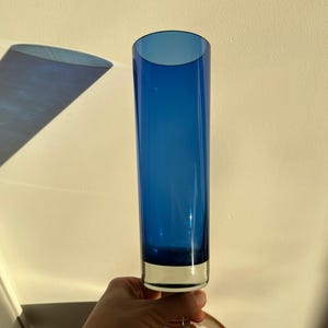 Op de afbeelding: Een hoge, kobaltblauwe glazen vaas met een heldere basis en steel. De vaas heeft een cilindrische vorm en wordt ondersteund door een heldere, cirkelvormige basis. De vaas is ongeveer 30 cm hoog.