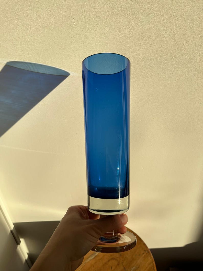 Op de afbeelding: Een hoge, kobaltblauwe glazen vaas met een heldere basis en steel. De vaas heeft een cilindrische vorm en wordt ondersteund door een heldere, cirkelvormige basis. De vaas is ongeveer 30 cm hoog.