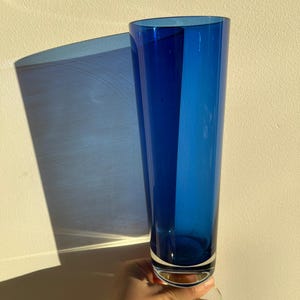 Op de afbeelding: Een hoge, levendige blauwe glazen vaas met een heldere, cirkelvormige basis. De vaas heeft een slanke, cilindrische vorm. De vaas wordt tegen een neutraal getinte muur gehouden, met een schaduw op de muur.