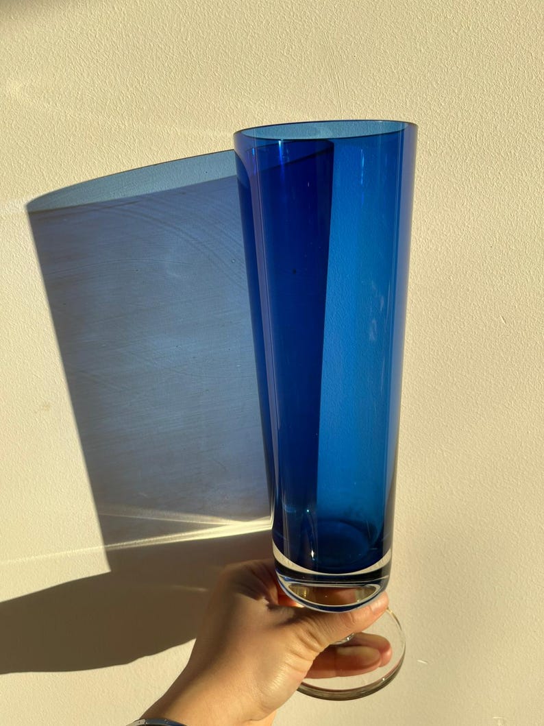Op de afbeelding: Een hoge, levendige blauwe glazen vaas met een heldere, cirkelvormige basis. De vaas heeft een slanke, cilindrische vorm. De vaas wordt tegen een neutraal getinte muur gehouden, met een schaduw op de muur.