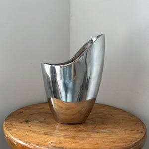 Pu&ograve; includere: Un vaso argentato lucido con un design unico e asimmetrico. Il vaso ha un'ampia apertura curva e una base arrotondata. La superficie riflettente del vaso crea un contrasto sorprendente con il tavolo di legno su cui poggia.