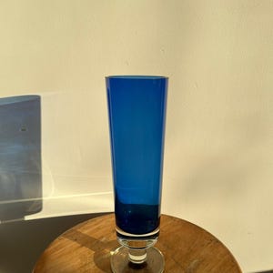 Op de afbeelding: Een hoge, kobaltblauwe glazen vaas met een heldere, cirkelvormige basis. De vaas heeft een slanke, cilindrische vorm en staat op een houten oppervlak. Het glas is diepblauw van kleur, de basis is helder.
