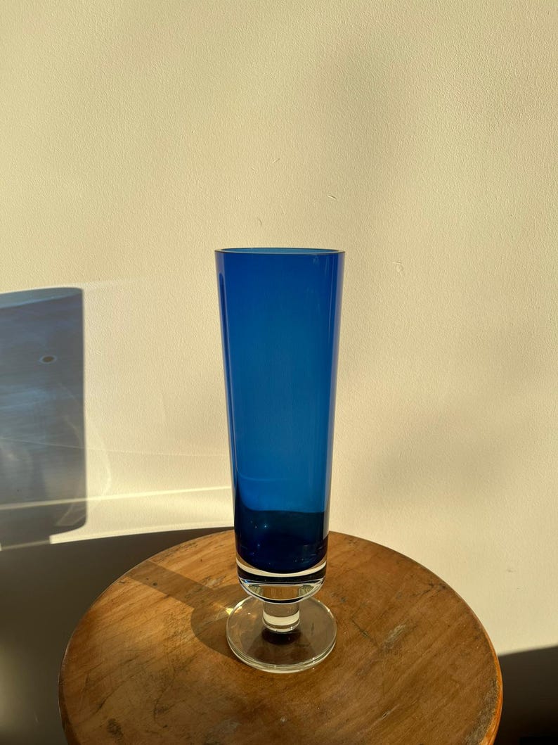 Op de afbeelding: Een hoge, kobaltblauwe glazen vaas met een heldere, cirkelvormige basis. De vaas heeft een slanke, cilindrische vorm en staat op een houten oppervlak. Het glas is diepblauw van kleur, de basis is helder.